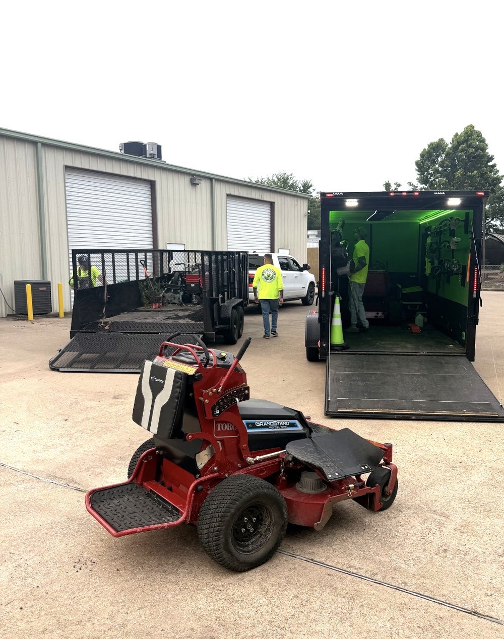 QuietScapes crew in hi-vis shirts loading the green-lit enclosed trailer, with a Toro GrandStand mower in front