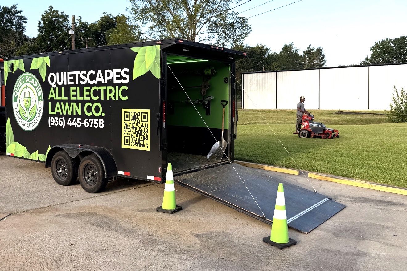 QuietScapes crew member mowing a commercial lawn beside the open QuietScapes All Electric Lawn Co. trailer, with safety cones at the ramp